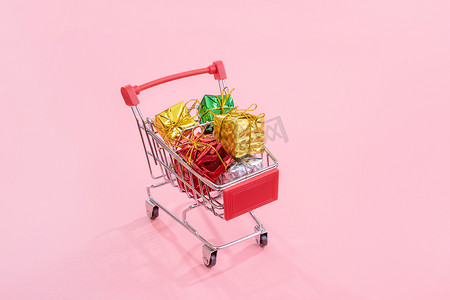 年度销售、圣诞购物季概念 — 迷你红色购物车手推车，里面装满了淡粉色背景中突显的礼盒，复制空间，特写