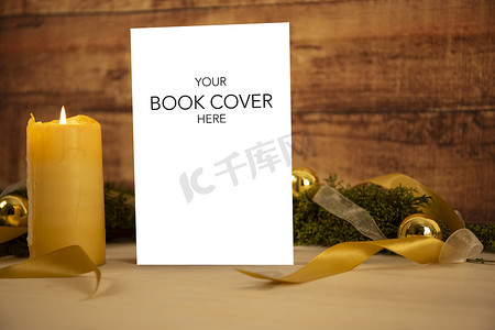 书籍展示的圣诞背景：空白封面的书上放着三支点燃的蜡烛、白色欧根纱和金色缎带、浅色木桌上的金色小玩意和深色木质背景