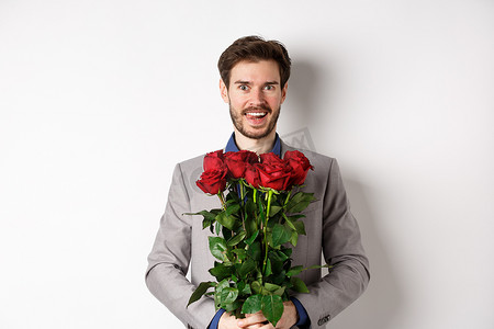 情人玫瑰摄影照片_穿着西装的帅哥兴奋地拿着一束玫瑰与情人浪漫约会，在情人节快乐地站着，白色背景