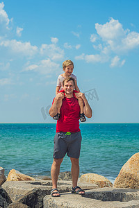 继父摄影照片_快乐的微笑父亲拥抱在肩膀上男孩男孩站在蓝色 lazur 海全景天际线地平线阳光明媚的天空。
