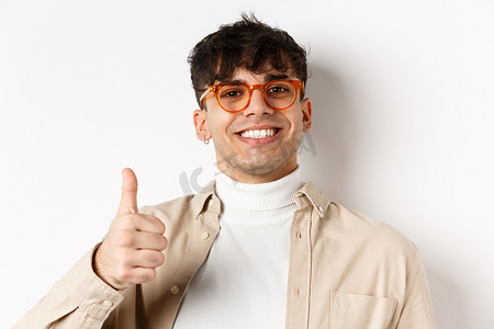 戴眼镜和耳环的帅气现代男人，满意地微笑着，竖起大拇指，推荐公司，站在白色背景上