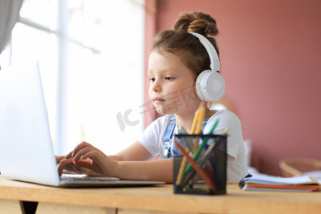戴着耳机的微笑小女孩在家里使用笔记本电脑在线学习手写，戴着耳机的可爱快乐的小孩在 PC 上上互联网课程或课程。