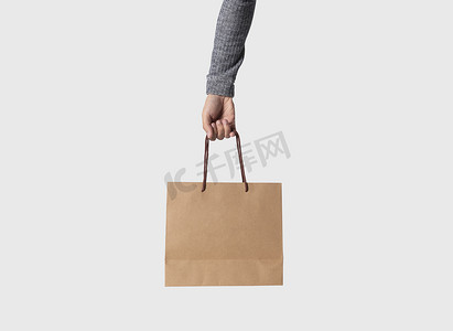 手持空白牛皮纸袋，用于在灰色背景上进行样机模板广告和品牌推广。