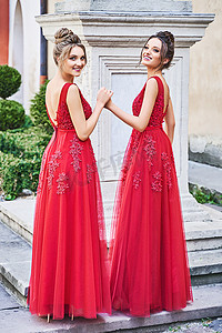 两个漂亮的伴娘女孩，金发女郎和黑发女郎穿着优雅的全长红色雪纺伴娘礼服，带蕾丝，手捧花束。