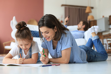 快乐的母亲微笑着女儿躺在温暖的地板上享受创造性活动，在相册中画铅笔着色图片，父亲躺在沙发上休息，家人一起度过空闲时间。