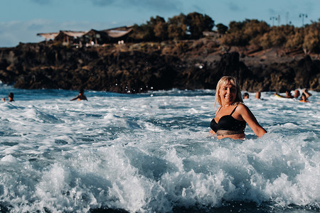 一个头发湿漉漉、穿着黑色泳衣的女孩站在特内里费岛的白色海水泡沫中，周围是浪花和水滴飞溅的海浪。加那利群岛。西班牙