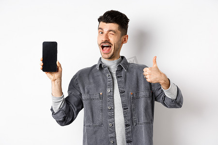 帅哥眨眼，用大拇指展示空手机屏幕，推荐应用程序或在线交易，站在白色背景下