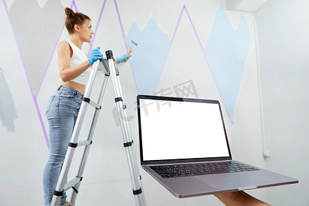 遮蔽胶带摄影照片_年轻女子用油漆滚筒刷墙，并使用遮蔽胶带站立 pn 梯子。