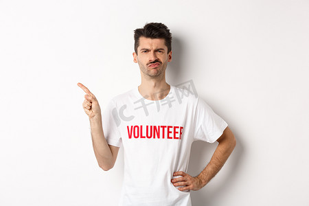 身穿 T 恤、持怀疑态度、犹豫不决的男性志愿者面带疑惑，指着手指离开促销优惠，白色背景