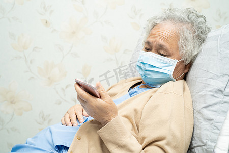 亚洲老年女性患者戴着面罩，在医院手持手机，以保护安全感染并杀死新型冠状病毒 Covid-19 病毒。