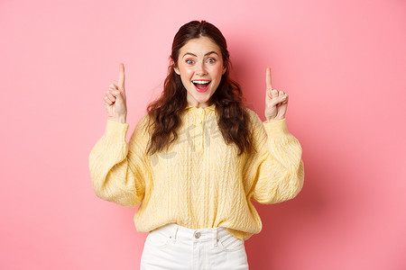 快乐的年轻女性展示广告，用手指指着促销文字，兴奋地微笑，站在粉色背景下