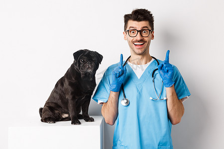 给人印象深刻摄影照片_兽医诊所英俊的年轻医生竖起手指，微笑着，站在可爱的黑哈巴狗旁边，白色背景，给人留下了深刻的印象
