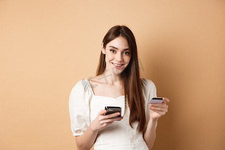 微笑的白人女性支付网上订单，拿着塑料信用卡和手机，无忧无虑地看着相机，米色背景