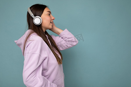 身穿浅紫色连帽衫、头戴白色蓝牙耳机的美丽微笑的黑发年轻女子的侧脸照片，她身穿浅紫色连帽衫，一边听着美妙的音乐，一边欣赏着