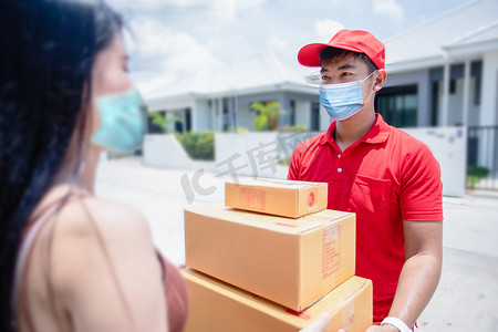 身穿红色制服、头戴红帽、戴着口罩的亚洲送货员正在搬运纸板箱，将其交给屋前的女顾客。