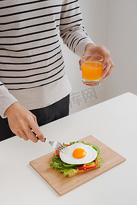鸡蛋汉堡摄影照片_人的手在纸上切鸡蛋，配上面包、香草、香肠和新鲜果汁。