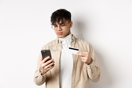 戴眼镜的帅气年轻人在电话上购物，在网上购物，拿着塑料信用卡和智能手机，站在白色背景上