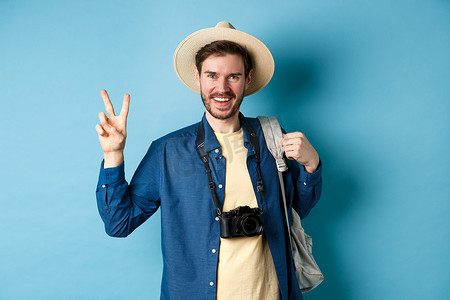 背包展示摄影照片_快乐帅哥暑假拍照，展示和平标志，微笑，戴着草帽，拿着旅游背包，蓝色背景
