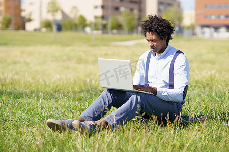 留着爆炸头的黑人男子坐在城市公园草地上的滑板上，使用他的笔记本电脑。
