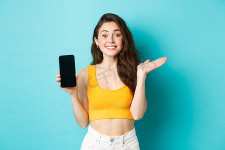 快乐的年轻女孩介绍您的应用程序或商店，展示空的智能手机屏幕，对着镜头微笑，站在蓝色背景下