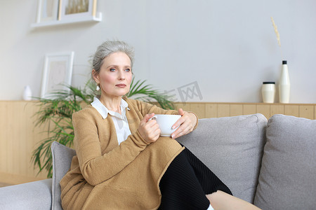 快乐的成熟女性躺在舒适的沙发上喝咖啡或茶，看着别处，在家里舒适的沙发上放松，享用热饮