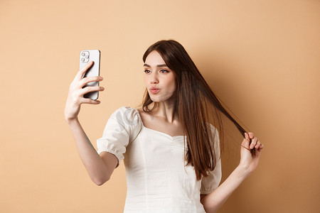 时尚的年轻女性玩头发，在智能手机上自拍，为社交媒体拍照，站在米色背景中