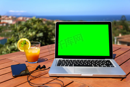 打开木桌上有绿色屏幕的笔记本电脑，配有智能手机和连接的耳机，以及背景中的蓝海全景，有选择地聚焦