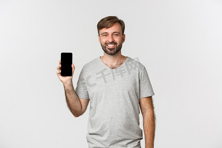 身穿灰色 T 恤、微笑帅哥的肖像，展示手机屏幕，推荐应用程序或购物网站，站在白色背景上