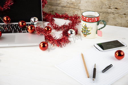 红色茶摄影照片_圣诞节假期的计算机工作概念：浅色木桌，配有开放式铝制笔记本电脑、红色装饰、红色小玩意、圣诞茶、智能手机、块笔记、铅笔和笔在块笔记上