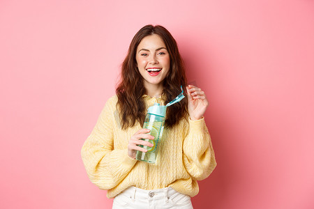 快乐的黑发女人的画像，头发卷曲，喝着私人瓶子里的柠檬水，笑着笑着，感觉健康，站在粉红色的背景下