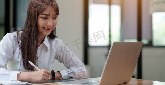 高管培训摄影照片_一个穿着白衬衫的年轻开朗的女孩微笑着，微笑着写下笔记，为学生在笔记本电脑桌面桌上进行高管培训