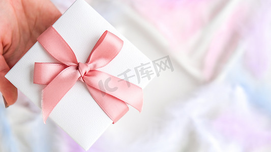 女手拿着白色礼品盒，白色丝绸纹理背景上有粉色丝带。