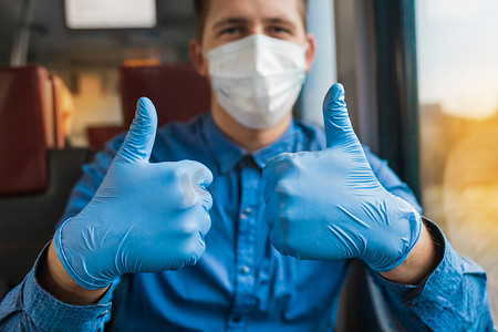 戴手套的护士摄影照片_戴着防护面具和医用蓝色手套的年轻白人男子竖起大拇指，特写