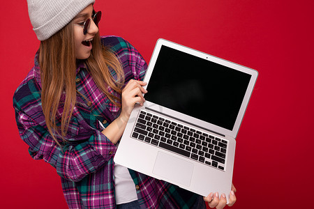 肖像照片中，美丽的金发年轻女子拿着电脑笔记本电脑，屏幕空空，戴着太阳镜帽子，穿着彩色衬衫，看着上网本显示屏和键盘，张开嘴，与红墙隔开，发出“哇”的肖像照片