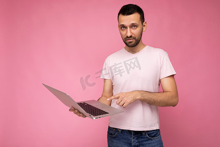 英俊的男人拿着笔记本电脑看着相机，指着孤立的粉红色背景中穿着 T 恤的屏幕