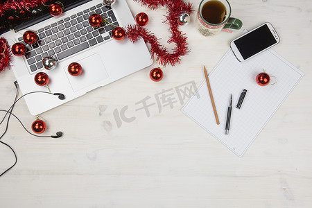 圣诞节假期的工作：平铺浅色木桌，配有开放式铝制笔记本电脑、红色装饰、红色小玩意、圣诞茶、智能手机、块笔记、铅笔和笔在块笔记上