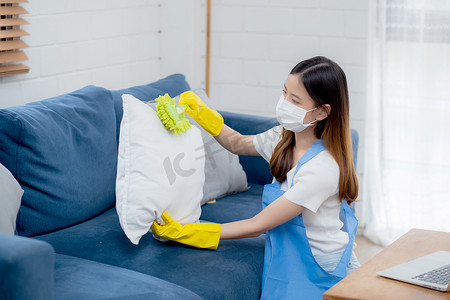 戴着面罩和手套的年轻亚洲女性在家里用沙发和垫子上的除尘器清洁灰尘，女孩在家里做家务以保持卫生、管家和服务、医疗保健和生活方式概念。