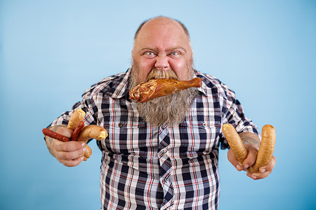 蓝色背景中，贪婪的肥胖男子用牙咬住鸡腿和成串的香肠
