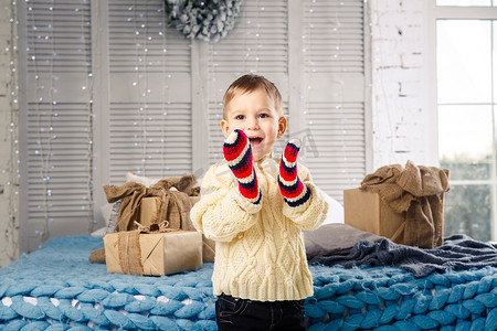哈哈大笑卡通人物摄影照片_圣诞节那天，一个有趣顽皮的小男孩坐在床上，手里拿着装着白色羊毛针织毛衣的礼盒，上面戴着明亮的大连指手套，大笑起来。