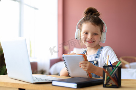 家庭教育家庭教育摄影照片_戴耳机的小女孩坐在办公桌前在笔记本上写字在线学习在家做运动，小孩子手写准备隔离作业，上网络课或在室内上课。