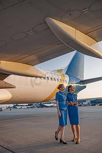 日落时，两名身着蓝色制服的空姐微笑着站在机场一架大型客机前的全长镜头