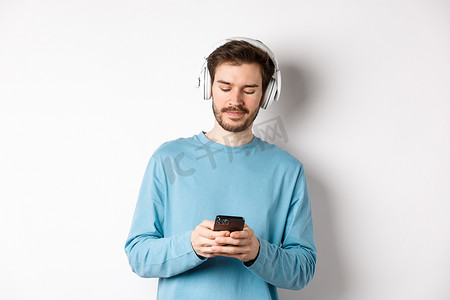 身穿蓝色运动衫的现代年轻人在智能手机上放音乐，用无线耳机听歌，白色背景