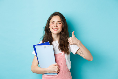 年轻女性上课，女学生学习，拿着笔记本，竖起大拇指表示赞同，推荐公司，站在蓝色背景上