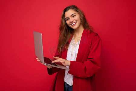 迷人微笑自信漂亮的年轻女士的侧面照片，手持笔记本电脑，身穿红色开衫和白色 T 恤，与红墙背景隔离。