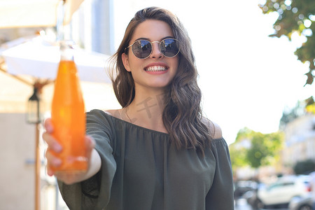 工作职责不清晰摄影照片_戴墨镜的年轻漂亮女人拿着一瓶果汁，看着相机，在户外。