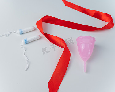 白色背景上的卫生棉条、粉色月经杯和红色缎带。