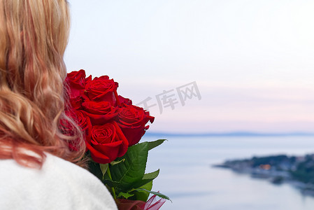 从酒店的海边阳台望去，身穿白色长袍、戴着一束红玫瑰的快乐女人