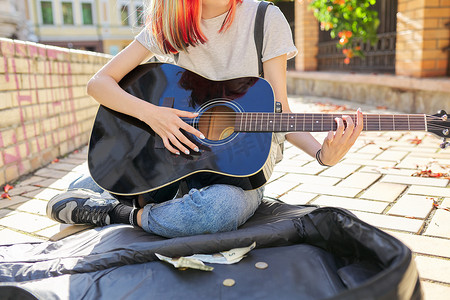 坐弹吉他摄影照片_弹吉他的创意女性坐在人行道上，带着吉他盒和现金