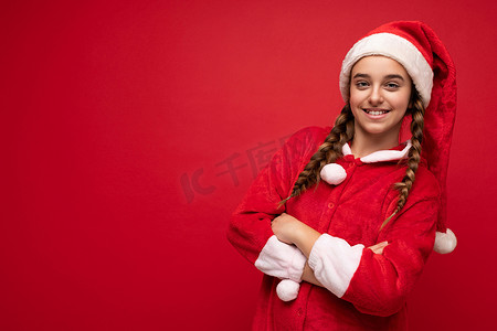 新年照片中，迷人迷人、令人惊叹、快乐、积极、微笑的黑发女孩，留着辫子，穿着圣诞老人套装，站在红色背景墙上，与世隔绝，看着相机