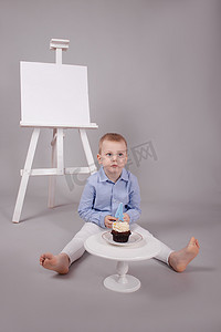 学龄前男孩穿着白色裤子和蓝色衬衫，灰色背景，靠近画架，有模型和带蜡烛的纸杯蛋糕，形状为四号 4。生日快乐。
